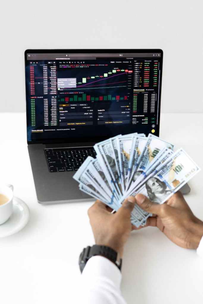 A person holding a fan of cash in one hand while looking at a laptop screen displaying stock market data.
