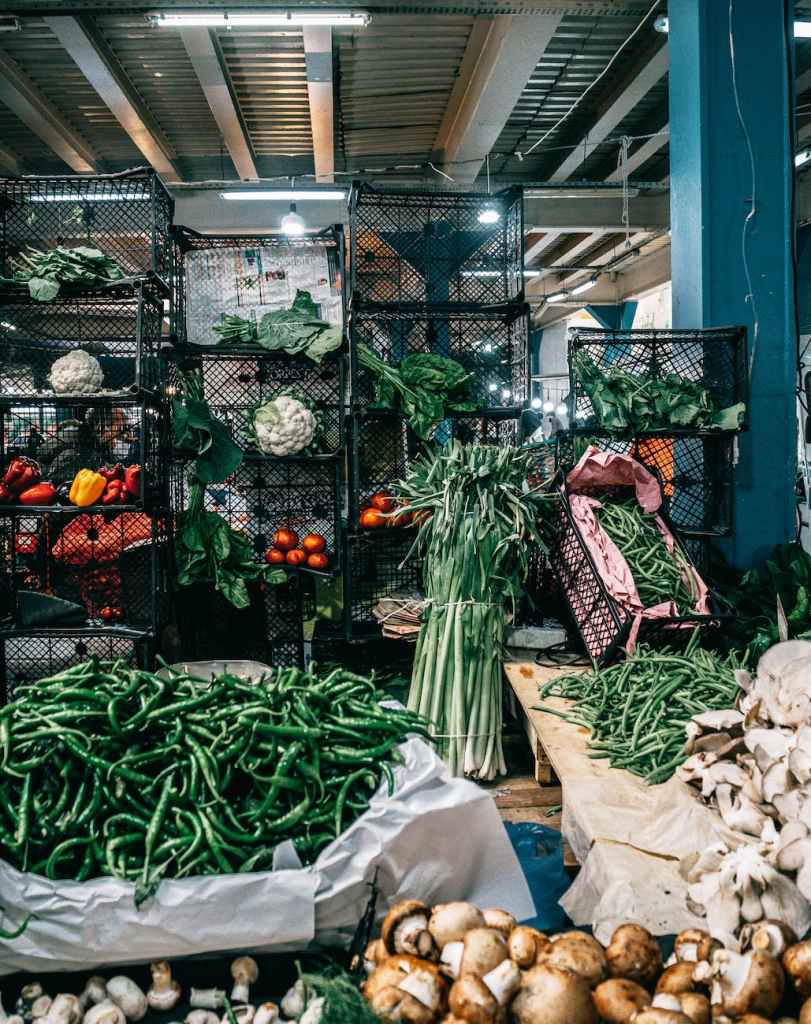 A vibrant display of fresh vegetables at a market, featuring green beans, mushrooms, tomatoes, and various leafy greens in wire baskets.