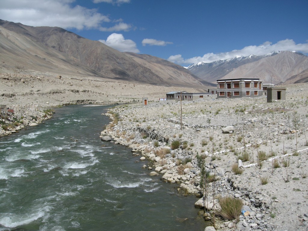 A picturesque view of a river flowing through a rocky landscape in Ladakh, with mountains in the background and a rustic building situated near the water.