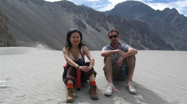 Two individuals sitting on a sandy slope in Nubra Valley, Ladakh, with mountains in the background.