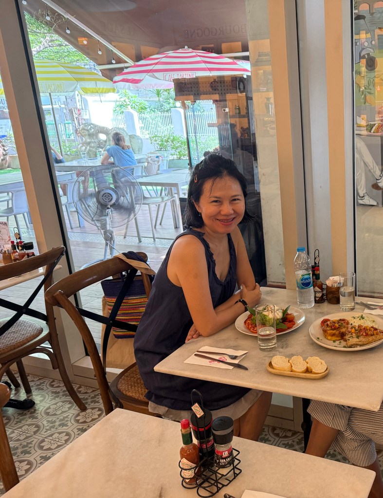 The author at a table in a restaurant, smiling while surrounded by food, drinks, and a window view of a patio with striped umbrellas.
