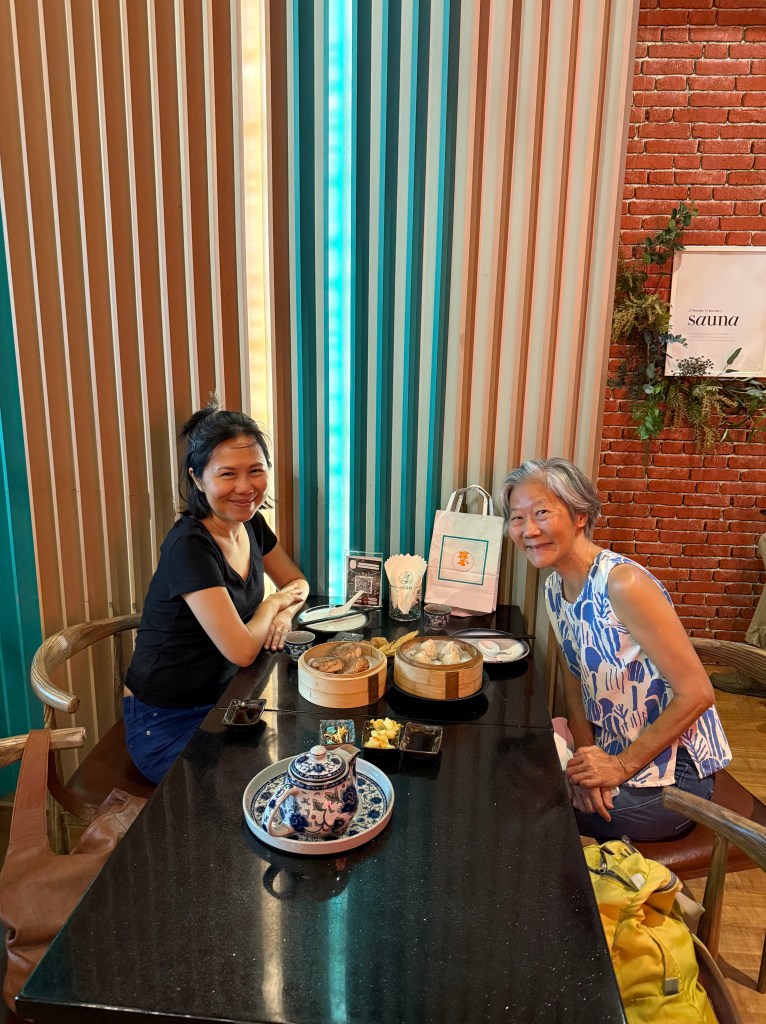 Two women sitting at a table with dim sum dishes and a teapot in a cozy restaurant, smiling and enjoying each other's company.