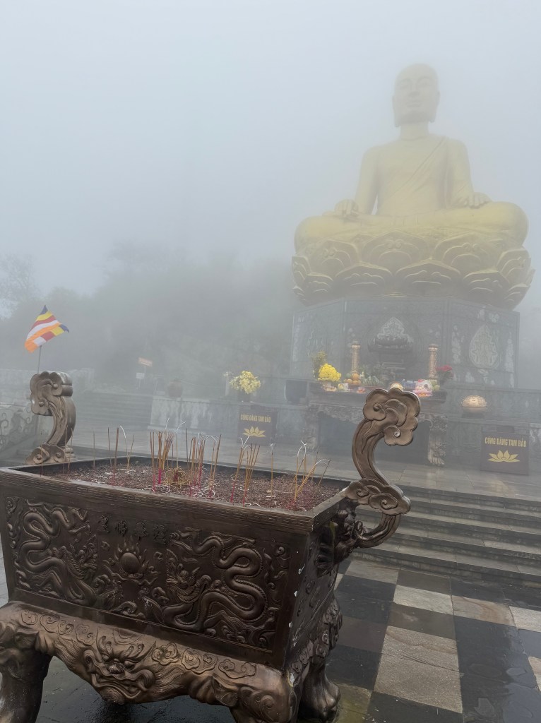 A misty scene at Yen Tu Mountain showing a large seated Buddha statue in the background, with incense burning in a decorative bronze urn in the foreground.