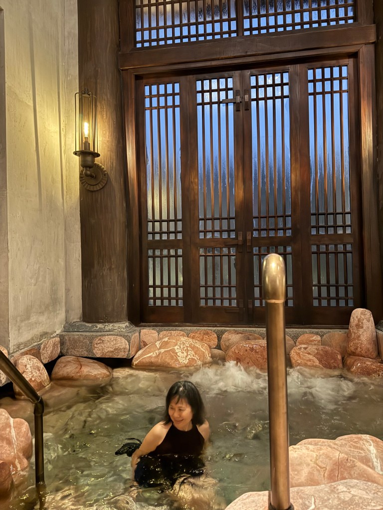A person relaxing in a mineral-rich hot spring, surrounded by stone and wooden decor, with soft lighting and frosted glass windows in the background.