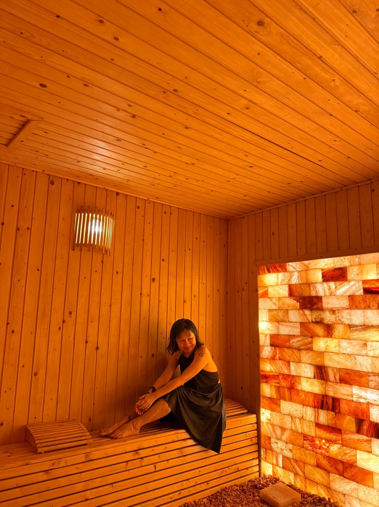 A woman sitting in a wooden sauna, featuring Himalayan  salt brick walls and warm lighting, creating a serene and relaxing atmosphere.