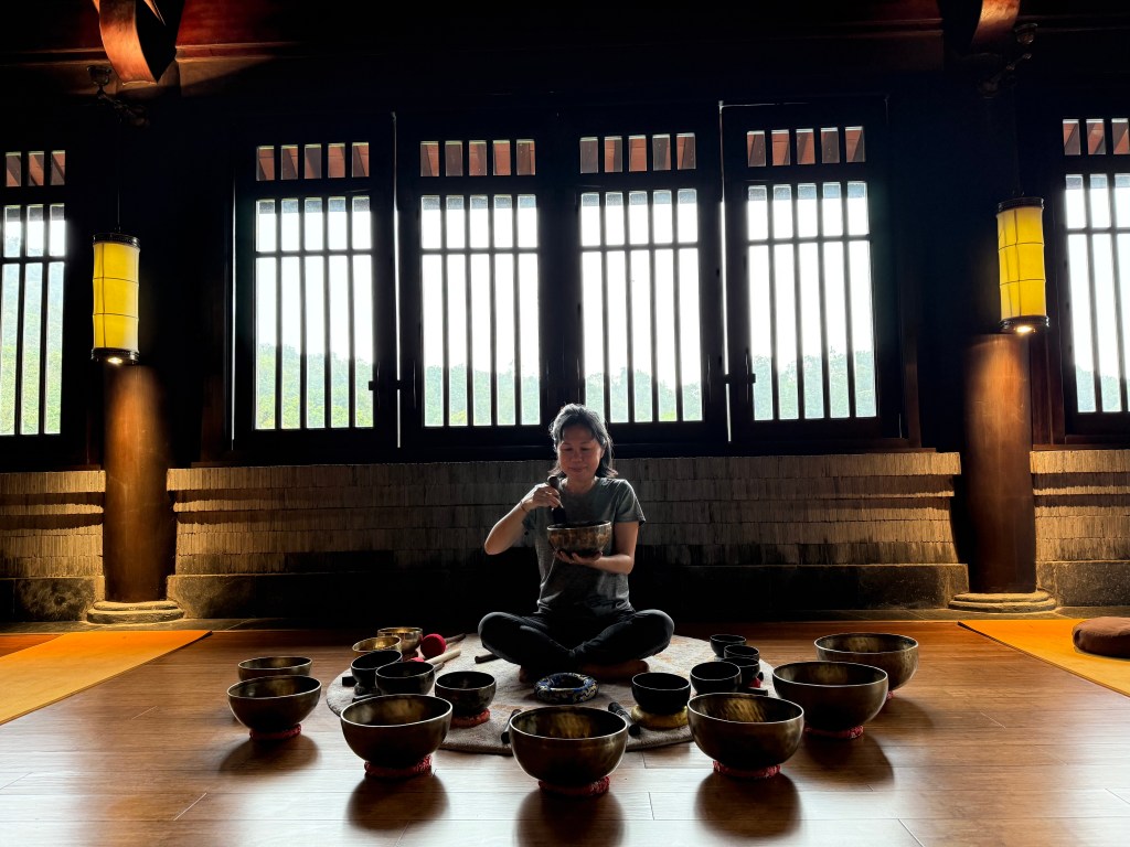 A person sitting cross-legged on a wooden floor, surrounded by various singing bowls, with sunlight streaming through large windows in the background.