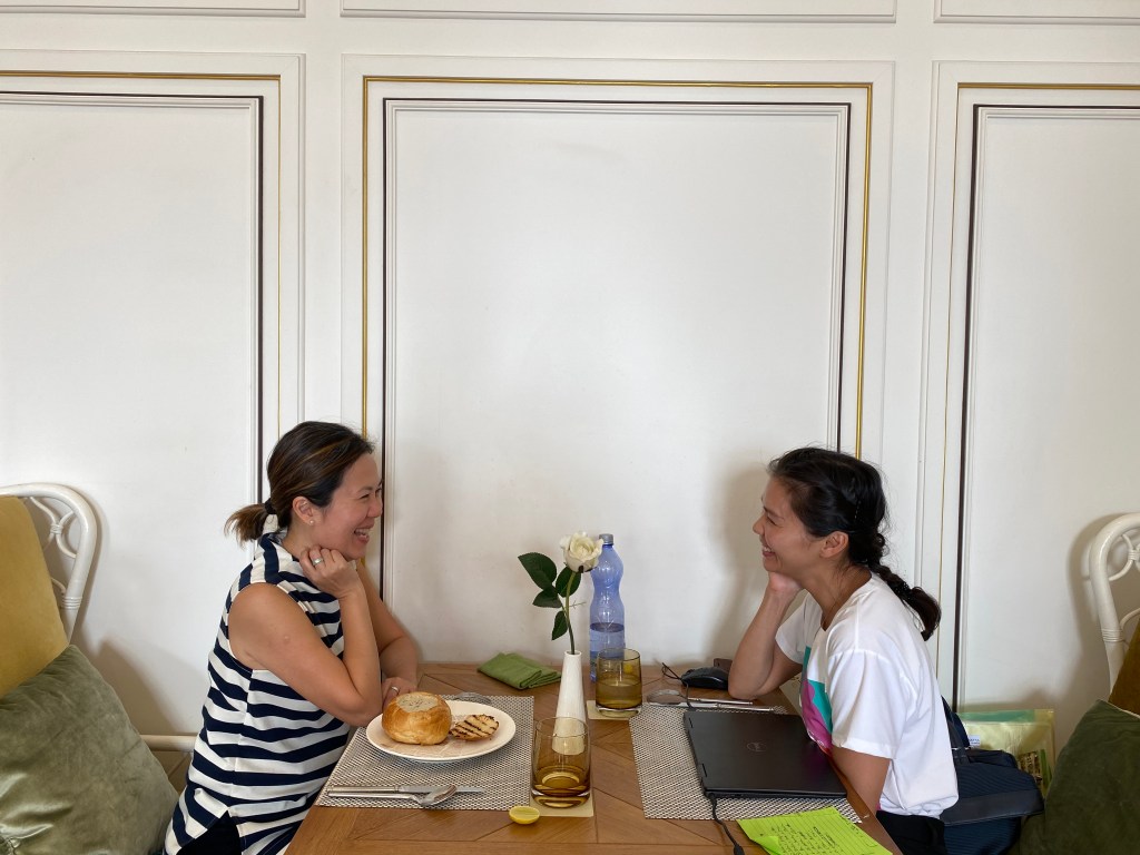 Two women sitting at a wooden table, engaged in a lively conversation over drinks and food, with a flower centerpiece and laptop beside them.