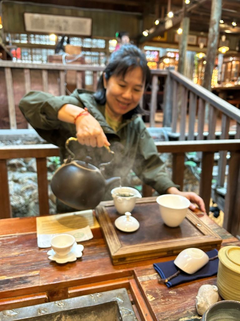 A woman pouring tea from a kettle into a small teacup at a traditional teahouse, surrounded by wooden decor and steaming tea.