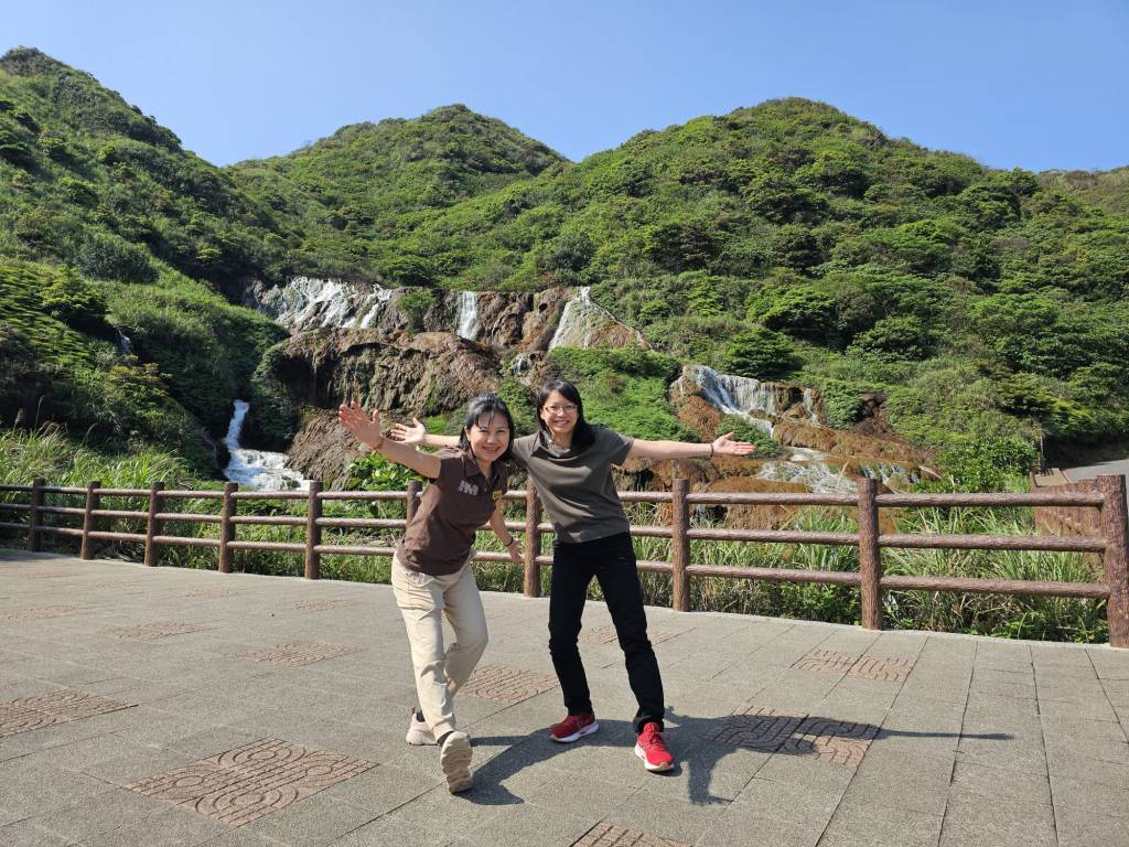 Two women posing joyfully in front of a lush green hillside with a waterfall in the background.