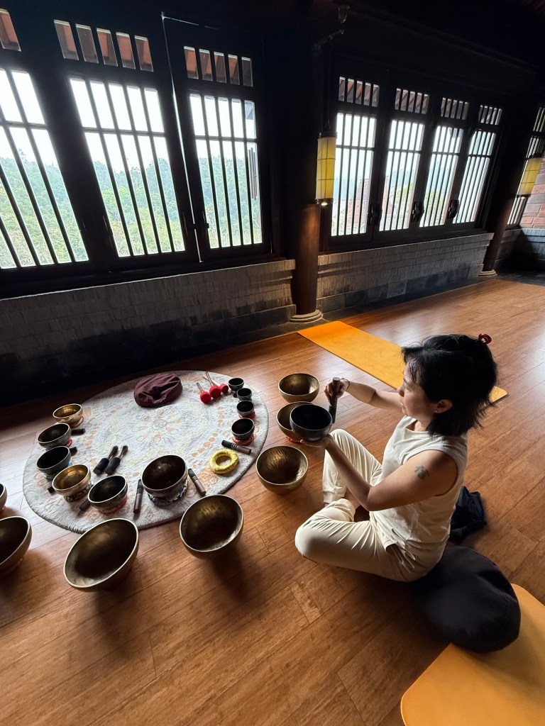 A woman sitting on a mat in a serene room, engaging with singing bowls arranged in a circular pattern on the floor, surrounded by natural light streaming through large windows.