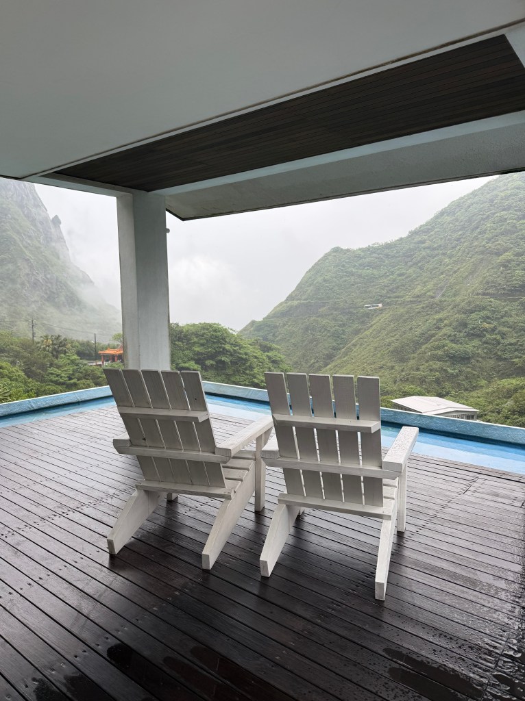 Two white adirondack chairs on a wooden deck with green mountainous scenery in the background under a cloudy sky.