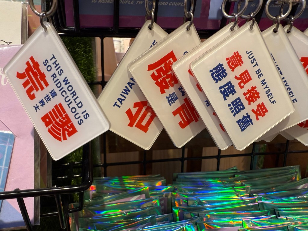 A collection of keychains displayed on a rack, featuring various phrases including 'THIS WORLD IS SO RIDICULOUS,' 'TAIWAN,' and 'JUST BE MYSELF' printed in both English and Chinese.