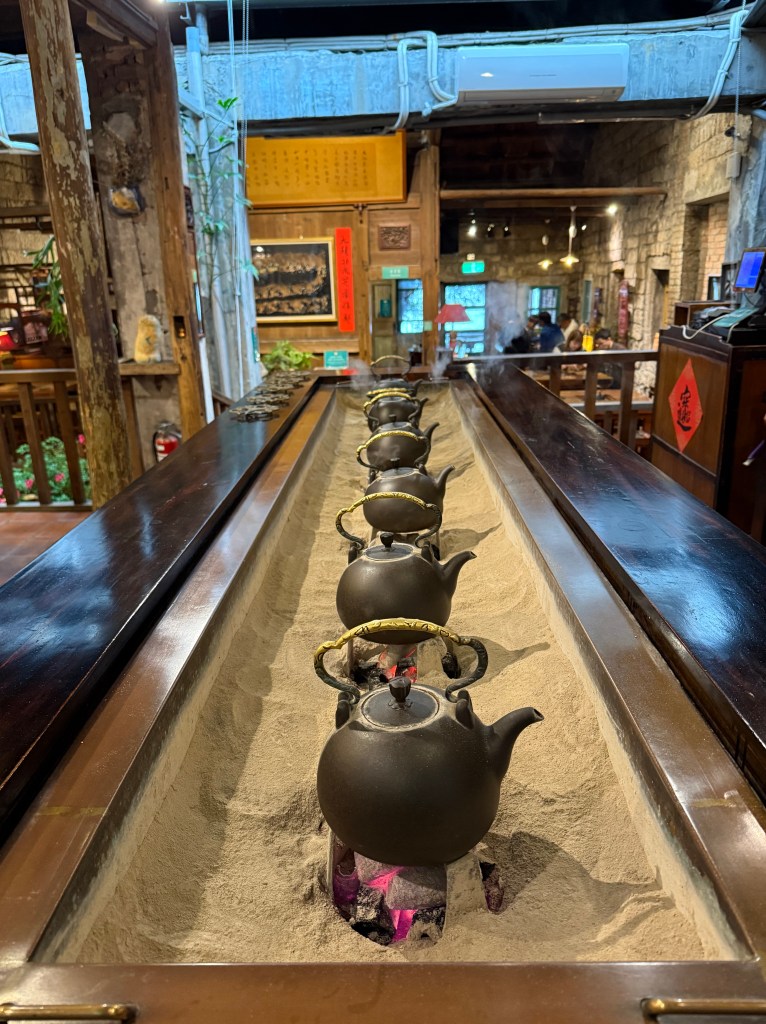 A cozy tea house interior featuring several black teapots placed in sand, with a rustic wooden table and traditional decor.