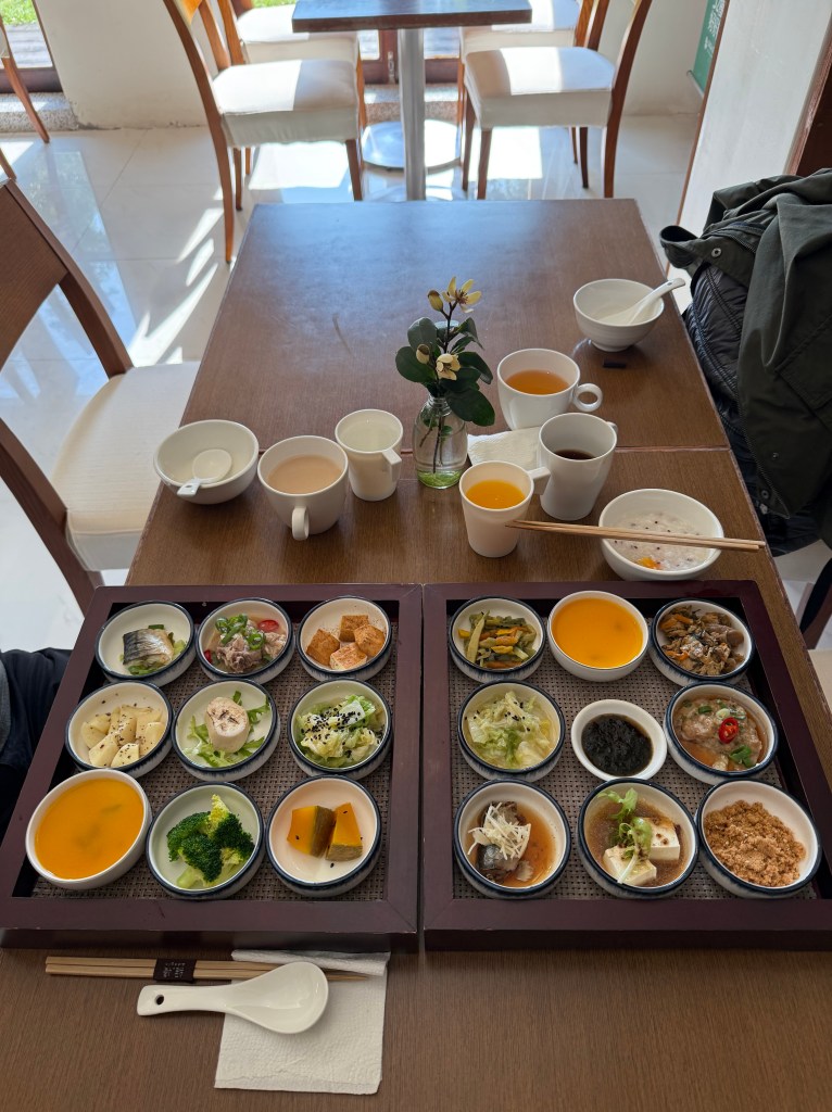 A beautifully arranged Chinese breakfast spread featuring small plates of various dishes including vegetables, tofu, and soups, accompanied by cups of tea and coffee, all set on a wooden table with a decorative flower.