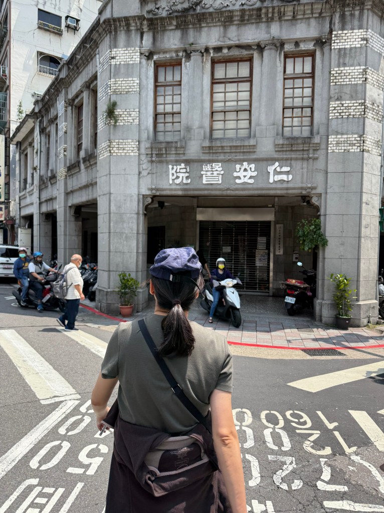 A person wearing a cap stands in front of a historic building with a decorative façade and Chinese characters. The street is lively with pedestrians and parked scooters.