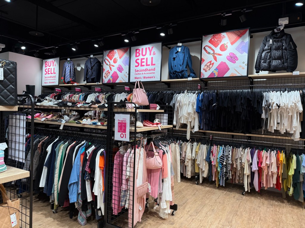 A retail store interior showcasing a variety of secondhand clothing and accessories, including men's and women's items, organized on racks and shelves. Signs above highlight the 'Buy & Sell' theme.