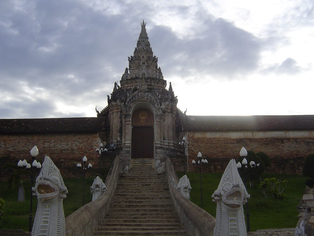 A traditional temple entrance with ornate architecture, flanked by stylized Nagas, leading up a staircase under a cloudy sky in Changmai.