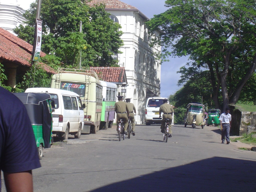 A street scene in Galle, Sri Lanka featuring two police officers riding bicycles, surrounded by parked vehicles including vans and tuk-tuks, with lush greenery and a colonial building in the background.