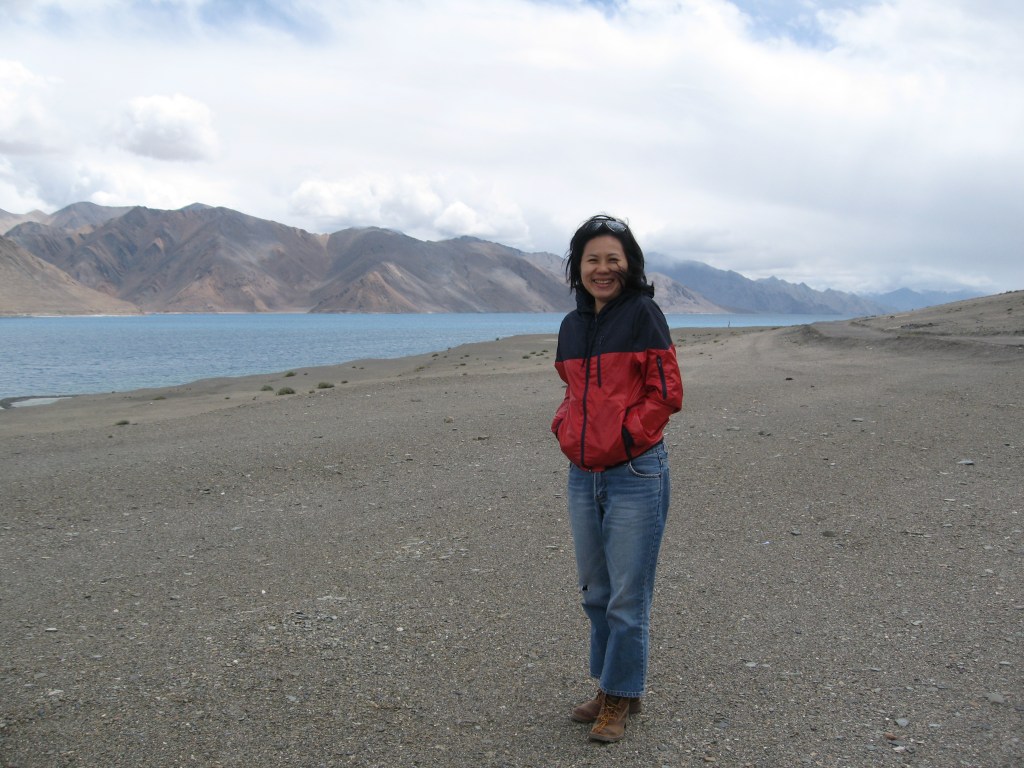 A person stands next  Pangong Tso, Ladakh with mountains in the background and a partly cloudy sky.