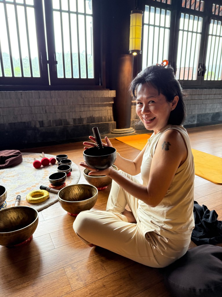 A person sitting cross-legged in a serene indoor setting, holding a small bowl and smiling, surrounded by various singing bowls and instruments.