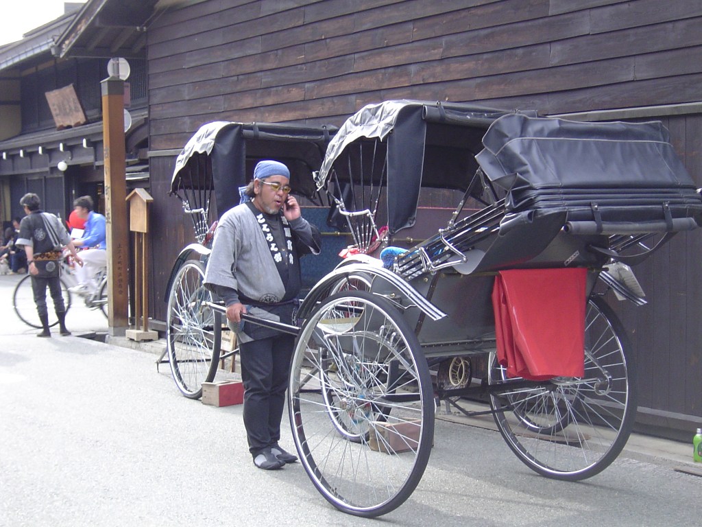A Japanese Rickshaw rider standing next to a traditional rickshaw, talking on a phone, in a street lined with wooden buildings.