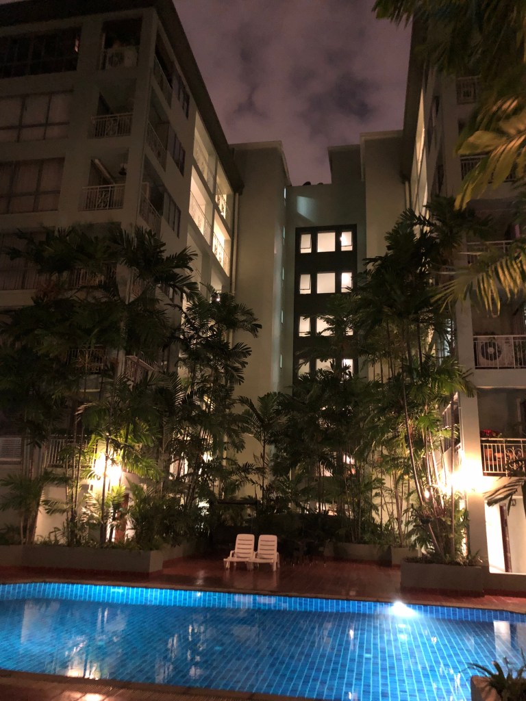 Night view of a residential building in Bangkok, showcasing a swimming pool surrounded by palm trees and illuminated by soft lighting.