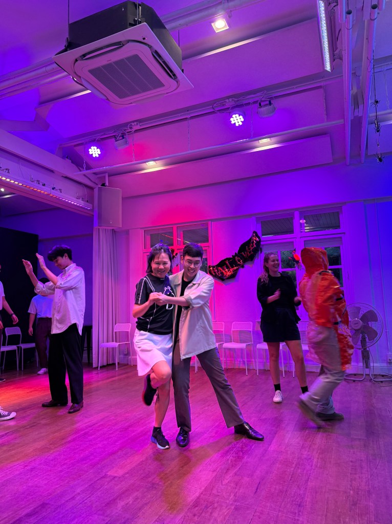 A group of dancers enjoying a lively swing dance session in a brightly lit studio, featuring vibrant colors and various dance styles. Two dancers in the foreground are striking a playful pose, showcasing enthusiasm and connection.