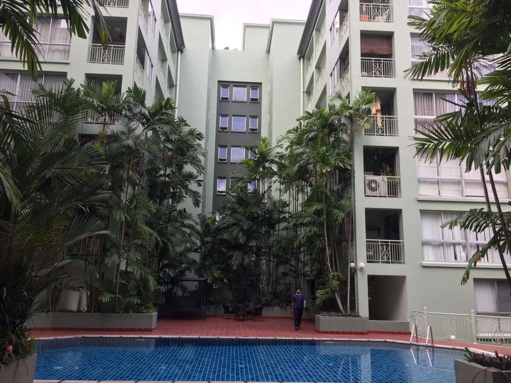 A view of a residential building in Bangkok surrounded by lush greenery and a swimming pool in the foreground.