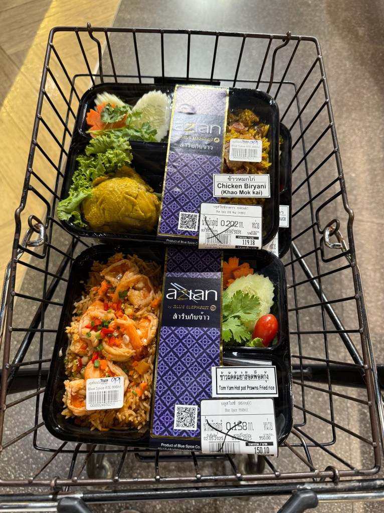 A shopping cart containing Thai food containers, including Chicken Biryani, Tom Yum Hot Pot Prawn Fried Rice, and Blue Spice Rice, alongside fresh vegetables.