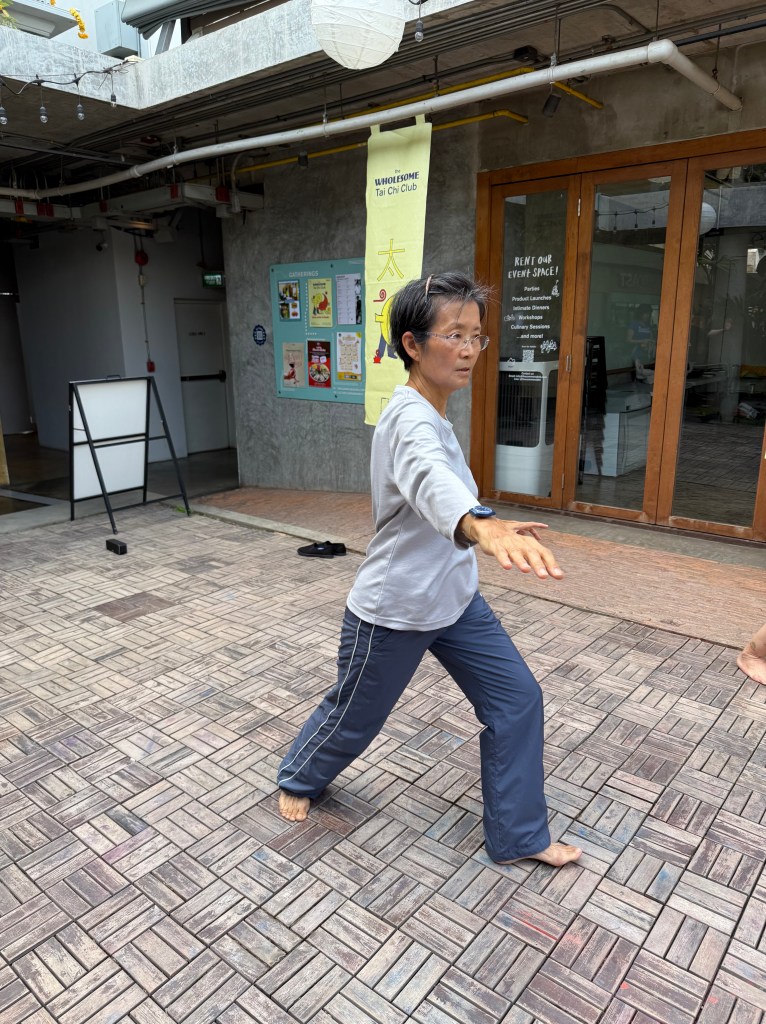 An older woman practicing Tai Chi in a peaceful outdoor setting at The Commons Thonglor, demonstrating a graceful movement with focused concentration.