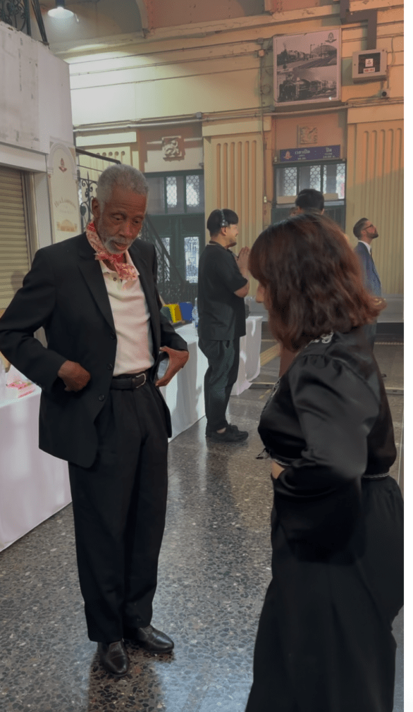 An older man in a black suit and red scarf interacts with a woman in a black dress at Hua Lumphong Station, surrounded by a lively atmosphere.