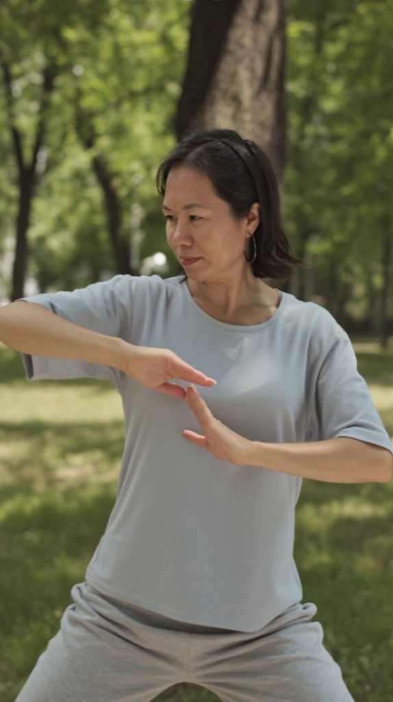 A woman practicing Tai Chi in a serene outdoor setting, wearing a light blue shirt and gray pants, demonstrating a flowing movement with focused expression.