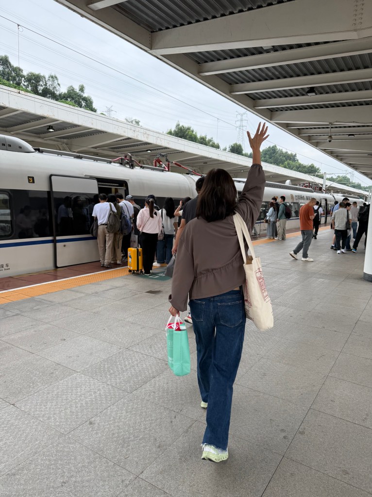 A traveler waving goodbye while standing at a train station in China, surrounded by other passengers and a high-speed train in the background.