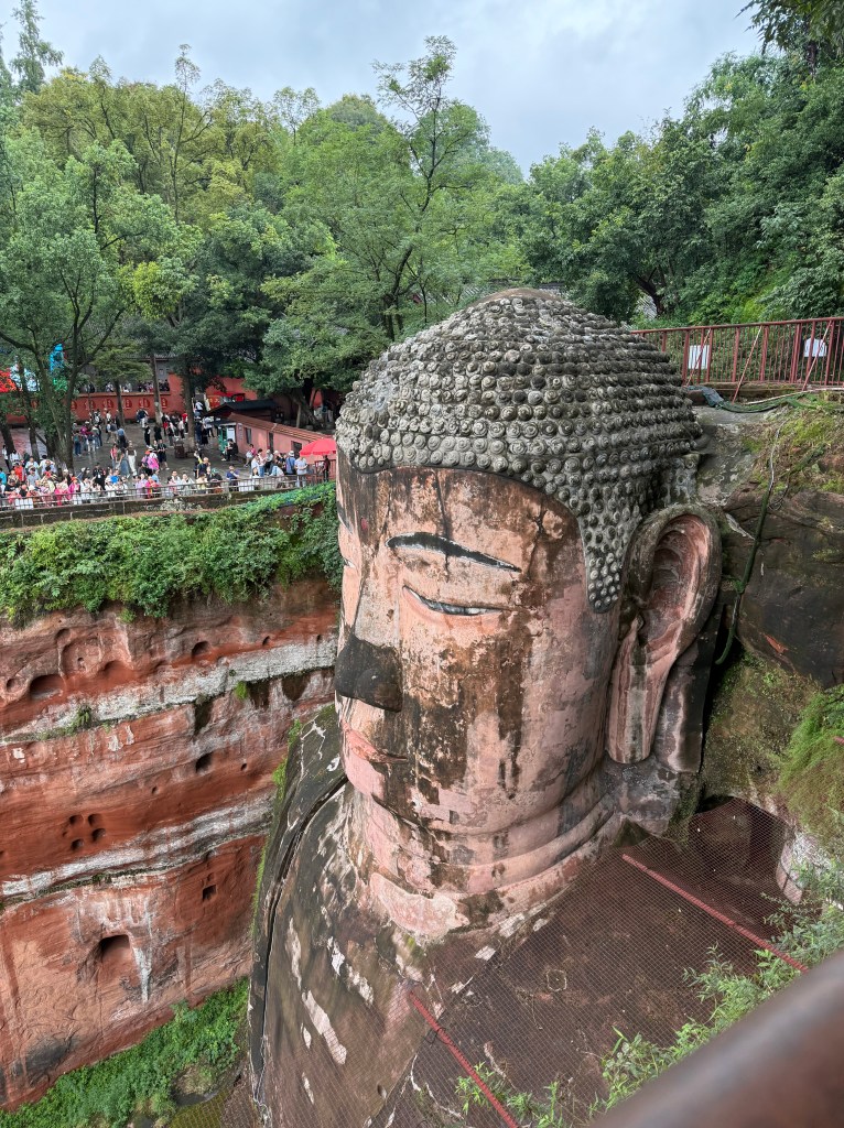 A close-up view of the Giant Buddha statue carved into a cliff, surrounded by lush greenery and visitors admiring the landmark.