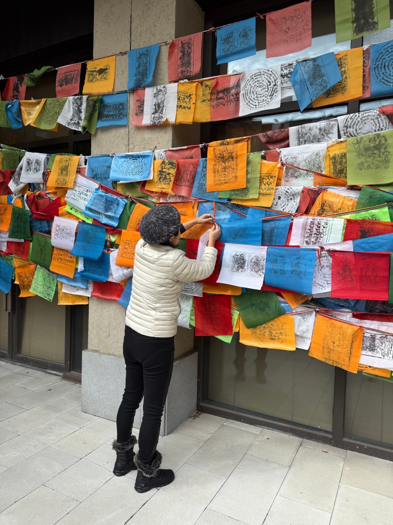 A person adjusting colorful prayer flags on a wall, wearing a light jacket and hat, with a serene expression.