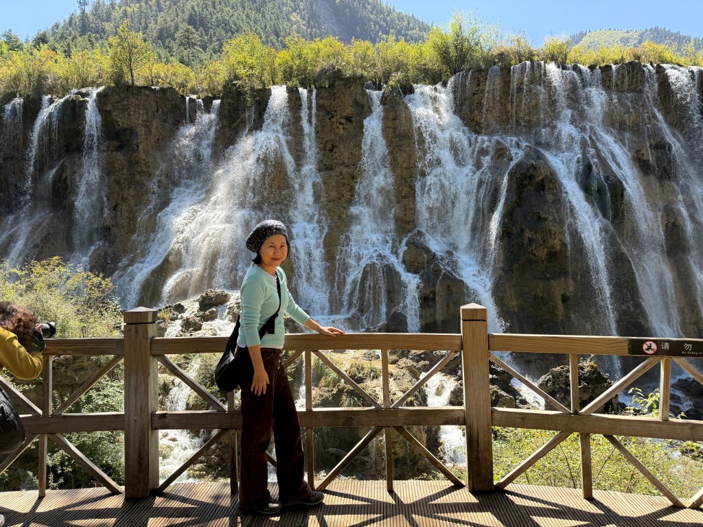 A woman stands on a wooden platform in front of a large waterfall, surrounded by trees and mountainous landscape.