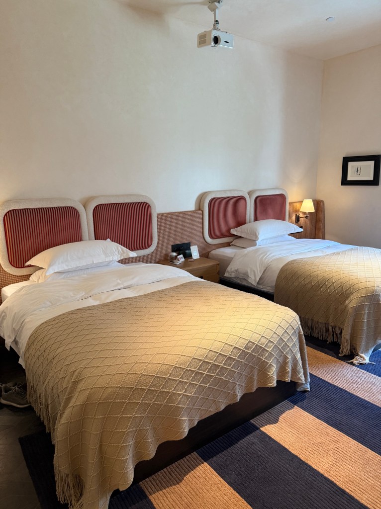 A cozy hotel room featuring two beds with beige woven blankets, a wooden bedside table, and a projector mounted on the ceiling.