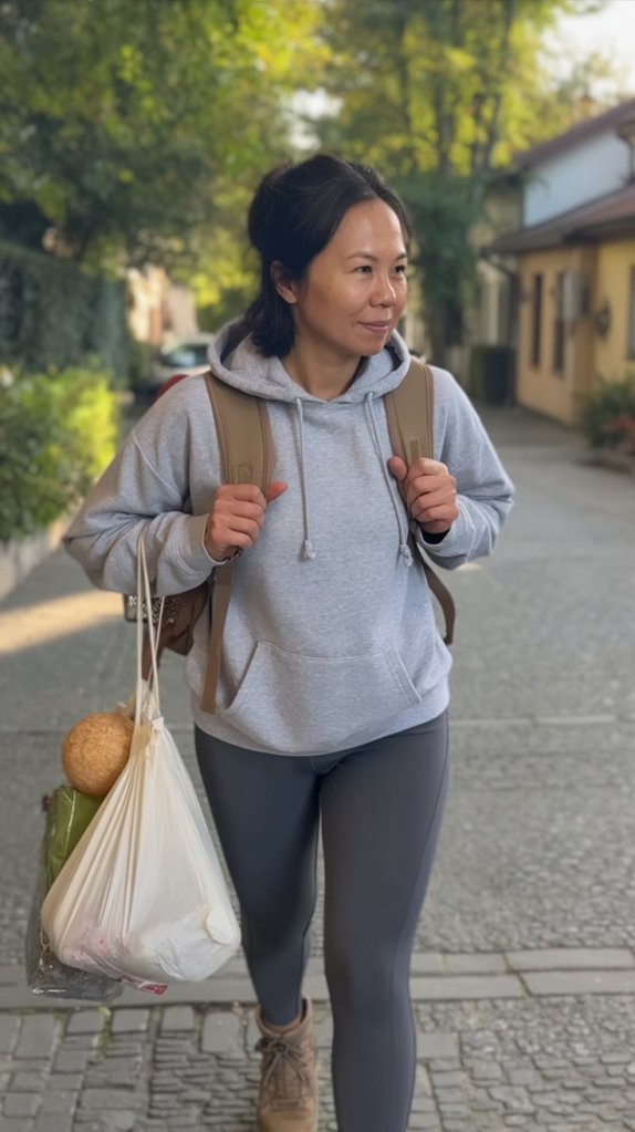 A person walking on a sidewalk, wearing a gray hoodie and leggings, carrying a backpack and a plastic bag filled with groceries.