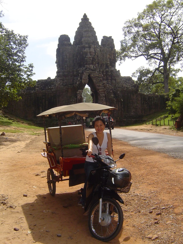 A woman sitting on a motorcycle next to a tuk-tuk in front of an ancient stone structure in Siem Reap, Cambodia.