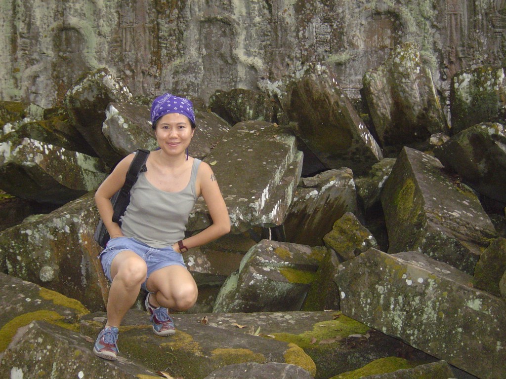 A woman crouches on moss-covered stones at an ancient temple, surrounded by large, weathered rocks, with a green landscape in the background.