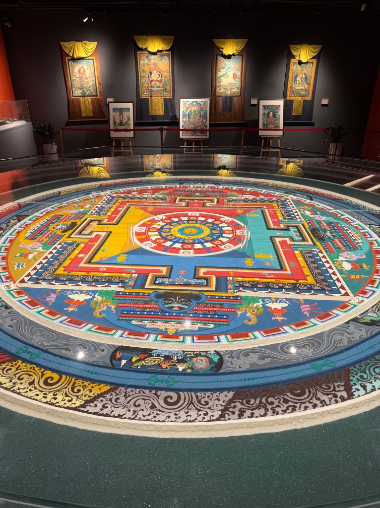 A colorful mandala displayed on the floor of a museum, surrounded by traditional Tibetan paintings hanging on the walls.