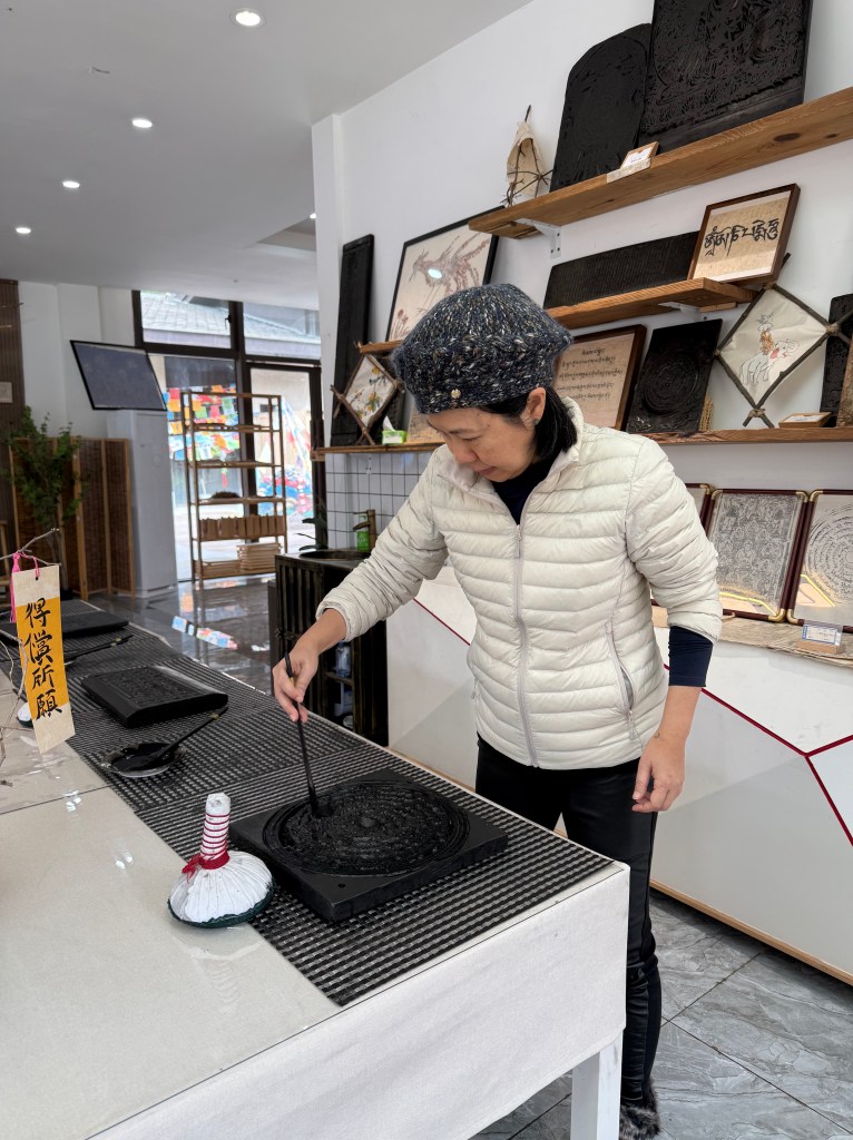 A woman in a cozy jacket is applying ink with a brush to a round stone plate in a craft workshop, surrounded by various traditional art pieces and materials.