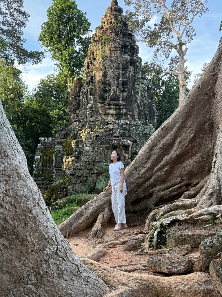 A person stands in front of a large stone monument with a serene face, surrounded by thick tree roots and lush greenery, capturing a moment of contemplation in nature.