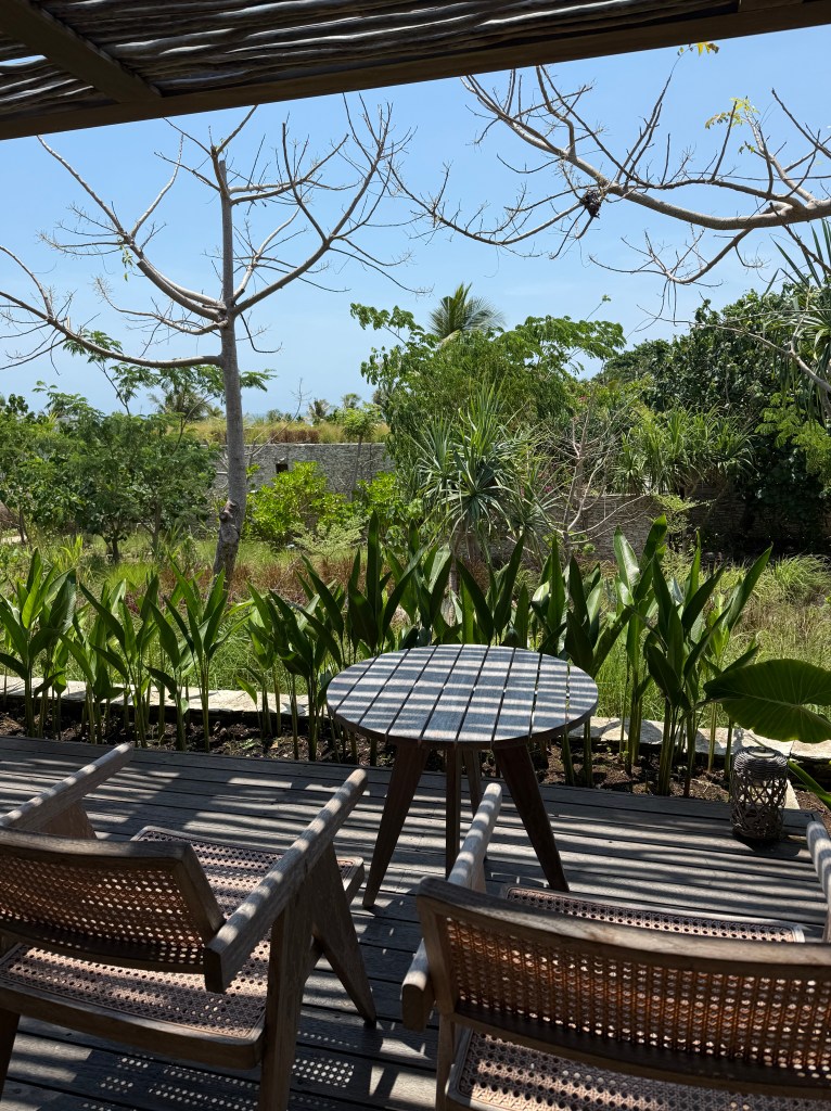 A peaceful outdoor seating area featuring two wooden chairs and a round table, surrounded by lush greenery and trees under a clear blue sky.