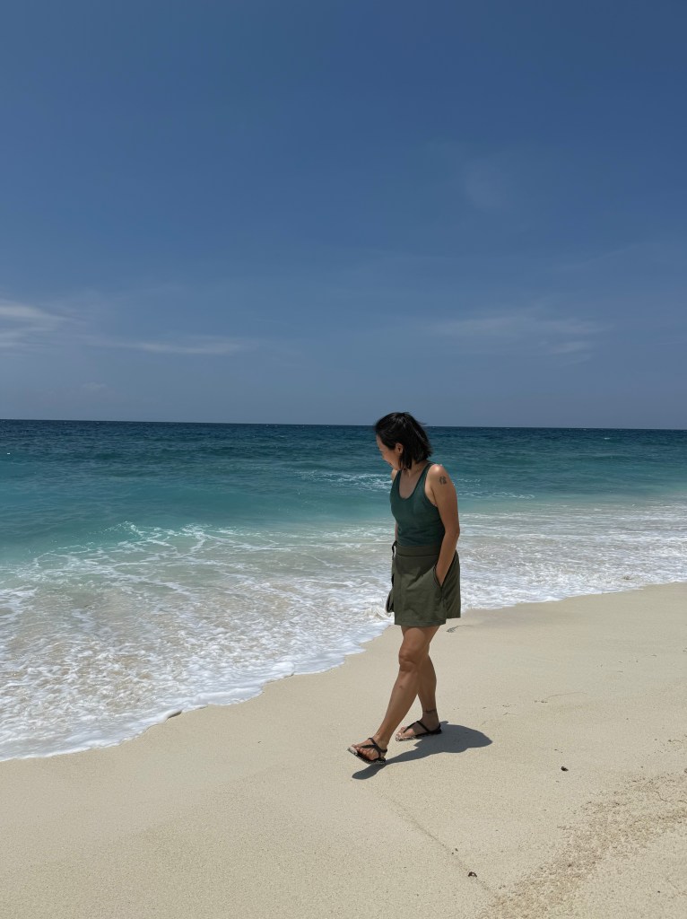 A person walking along a sandy beach with gentle waves lapping at the shore under a clear blue sky.