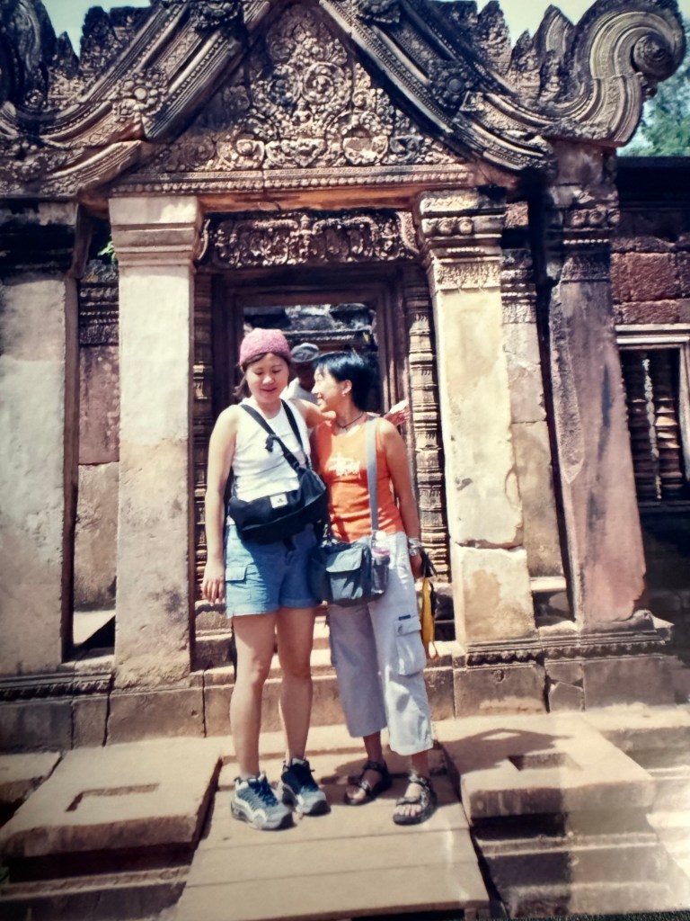 Two women smiling and posing together in front of a beautifully carved stone structure, likely part of an ancient temple.