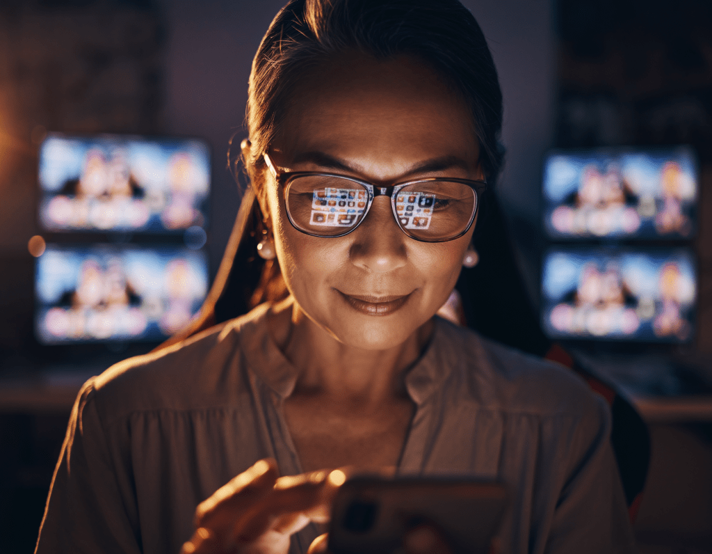 An older woman wearing glasses looks at her smartphone, with reflections of images from computer screens behind her, set in a softly lit room.