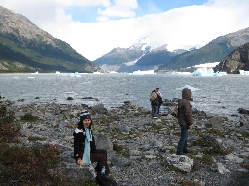 A person sitting on a rocky shore beside a glacial lake, with mountains and icebergs in the background. Two other people are walking along the shore, exploring the scenic landscape.