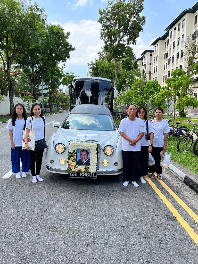 A group of mourners in white attire stands next to a flower-adorned car with a framed photo in front, parked on a street lined with trees and apartment buildings.