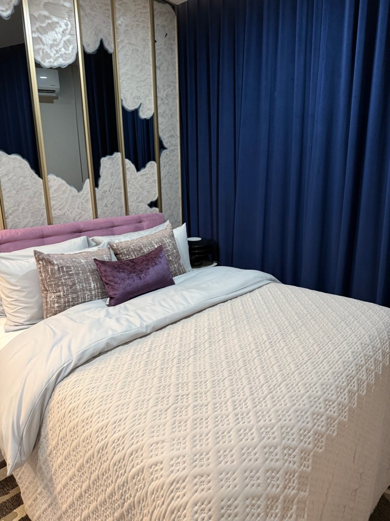 A modern, well-decorated bedroom featuring a neatly made bed with a textured white duvet and decorative pillows, set against a backdrop of dark blue curtains and a mirrored wall.